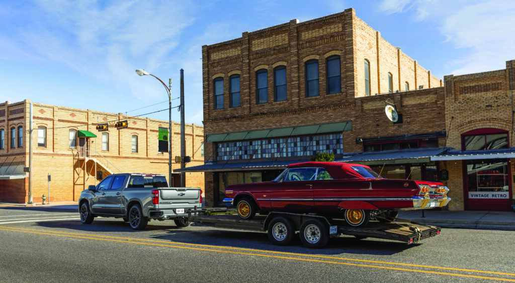 Modern Chevrolet Silverado towing a classic American lowrider on a trailer through a historic downtown street.