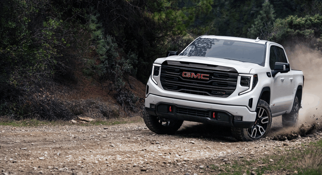 2026 GMC Sierra 1500 AT4 driving on a gravel road through a wooded landscape.