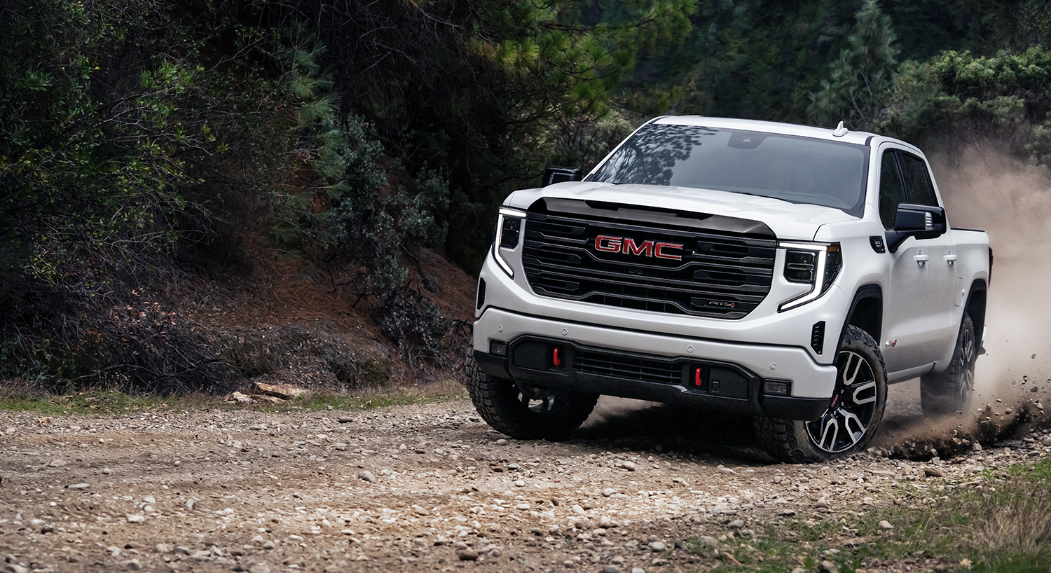 2026 GMC Sierra 1500 AT4 driving on a gravel road through a wooded landscape.